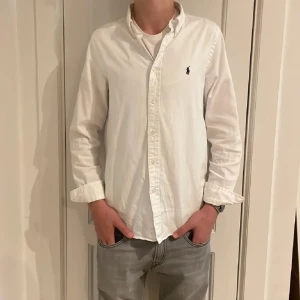Vit skjorta från Ralph Lauren - Säljer en klassisk vit skjorta från Ralph Lauren med den ikoniska loggan broderad på bröstet. Skjortan har långa ärmar och knappar framtill. Perfekt för en stilren look. storleken är 18år men passar xs/s