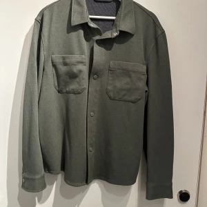 Grön overshirt med tryckknappar - Säljer en stilren grön overshirt med tryckknappar och två stora bröstfickor. Perfekt för lager-på-lager-stil. Den har långa ärmar och en klassisk krage.