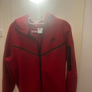  Nike - Säljer en snygg röd hoodie från Nike med dragkedja och huva. Den har aldrig varit använd förut och har även byxorna om man är intresserad ! Skick 10/10 