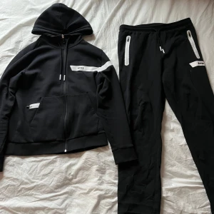 Svart tracksuit från boss  - Säljer en stilren svart tracksuit från BOSS med dragkedja och vita detaljer. Hoodien har en justerbar huva och praktiska fickor framtill. Perfekt för en avslappnad stil. Nypris 3298:- SEK 
