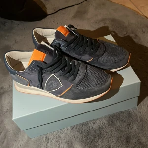 Mörk Blå sneakers ifrån Philippe Model - Snygga grå sneakers från Philippe Model med orange detaljer. Skorna har en kombination av mocka och meshmaterial, vilket ger en stilren look. De har snörning och en bekväm sula. Perfekta för kyla, springa, och värme. In princip helt nya, priset går att diskutera