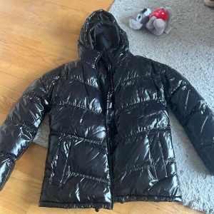 Svart pufferjacka med huva - Snygg svart pufferjacka med glansig yta och huva. Jackan har dragkedja framtill och två fickor på sidorna. Perfekt för kyliga dagar.