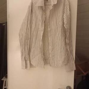 Vit och svart  och brun randig skjorta - Snygg vit skjorta med svarta ränder. Den har långa ärmar och knappar framtill. Perfekt för en stilren look. Stl s