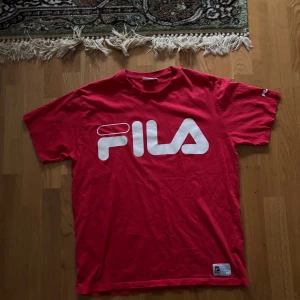 Röd t-shirt från FILA - Säljer en röd t-shirt från FILA med stort vitt logotryck på framsidan. T-shirten har korta ärmar och en klassisk rund halsringning. Perfekt för en sportig look eller avslappnad stil.
