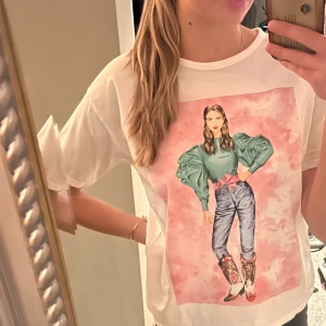 Vit t-shirt med tryck från Zara - Säljer en vit t-shirt från Zara med ett färgglatt tryck av en kvinna i grönt och rosa på framsidan. T-shirten har en rund hals och korta ärmar. Perfekt för en avslappnad stil.