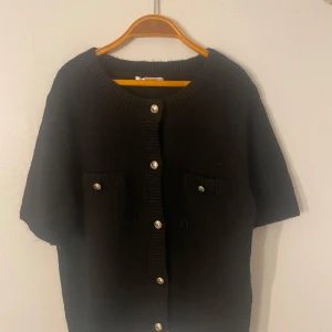 Svart kofta från Gina Tricot - Säljer en stilren svart kofta från Gina Tricot med korta ärmar och knappar framtill. Koftan har två dekorativa fickor med knappar och en ribbad kant vid halsen. Perfekt för en elegant look.