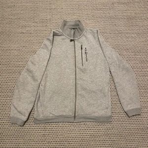 Grå ziphoodie från Sail Racing - Snygg grå ziphoodie från Sail Racing med dragkedja och en praktisk bröstficka. Perfekt för en stilren look. Jackan har en hög krage och är långärmad, vilket gör den idealisk för kyligare dagar. Fantastiskt bra skick!