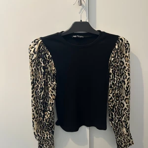 Svart topp med leopardmönstrade ärmar från Zara - Säljer en cool svart topp från Zara med leopardmönstrade puffärmar. Toppen har en rund hals och är perfekt för att ge en trendig touch till din outfit. Ärmarna har knappar vid ärmsluten för extra stil. plus har aldrig använt denna så den är helt oanvänd 💕