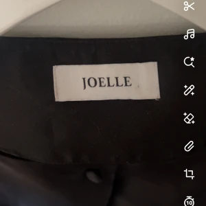 Svart kavaj från Joelle - Snygg svart kavaj från Joelle med puffiga ärmar och knappar framtill. Perfekt för en stilren look. Kavajen har en elegant design och är tillverkad i ett glansigt material som ger en lyxig känsla. Använd 2 gånger 