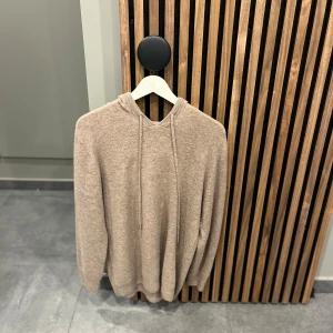 Beige stickad hoodie - Säljer en beige merinoull hoodie med långa ärmar och justerbar huva. Perfekt för kyliga dagar med sin mjuka och bekväma passform. Ribbstickade muddar vid ärmslut och nederkant ger en stilren look. Priset sitter som berget.
