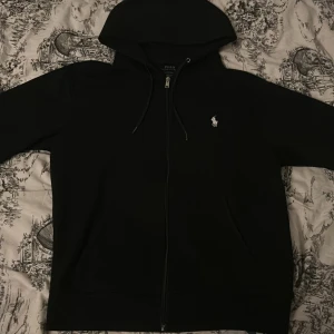 Svart hoodie från Ralph Lauren - Säljer en stilren svart hoodie från Ralph Lauren med dragkedja och det klassiska logotypen på bröstet. Perfekt för en avslappnad look. Den har en bekväm passform och huva med dragsko.