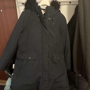 Svart parkajacka från GANT - Säljer en stilren svart parkajacka från GANT med pälskantad huva och dragkedja. Jackan har långa ärmar och praktiska fickor med knappar. Perfekt för kyliga dagar. Sparsamt använt marinblå/navy med huva och avtagbar pälskant. Storlek XL, jackan har flera fickor. Nypris ca 4499kr