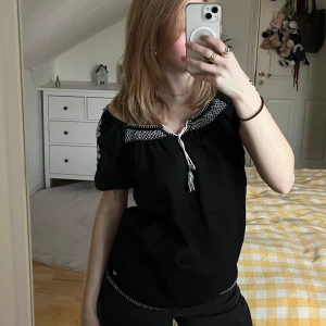 Svart offshoulder blus med broderi - Snygg svart offshoulder blus med vita broderade detaljer runt halsen och ärmarna. Blusen har en knytning framtill som ger en bohemisk känsla. Perfekt för en avslappnad stil.
