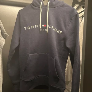 Mörkblå hoodie från Tommy Hilfiger - Tja! Säljer en snygg mörkblå hoodie från Tommy Hilfiger i ett väldigt bra skick då den aldrig är använd. Med vit logga på bröstet och känguruficka. Hör av dig vid funderingar!