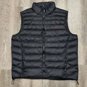 Svart dunväst från Polo Ralph Lauren - Snygg svart dunväst från Polo Ralph Lauren med röd broderad logga på bröstet. Västen har dragkedja framtill och är perfekt för kyligare dagar. Den quiltade designen ger en stilren look.