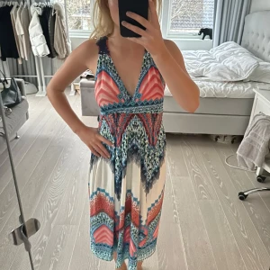 Färgglad klänning från Do Do - Säljer en färgglad klänning från Do Do med ett unikt mönster i blått, rött och vitt. Klänningen har en v-ringad design och spetsdetaljer vid axlarna. Perfekt för sommarens äventyr!  Storlek M/L men det är italiensk storlek så det är en S/M. 