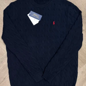 Mörkblå stickad tröja från Ralph Lauren - Säljer en stilren mörkblå stickad tröja från Ralph Lauren med det klassiska röda broderade logotypen på bröstet. Den är helt ny och prislapp ingår. Tröjan har en rund halsringning och långa ärmar, perfekt för kyligare dagar. Passar både till jeans och chinos för en avslappnad look. Kontakta gärna för frågor eller prisförslag.