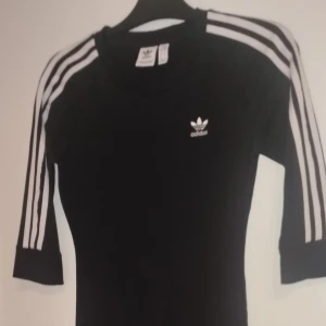 Svart klänning från Adidas - Snygg svart klänning från Adidas med de ikoniska tre ränderna på ärmarna. Klänningen har en normal passform och trekvartsärmar. Perfekt för en sportig och stilren look.