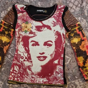 Marilyn monroe tröja desigual - Unik långärmad topp från Desigual med, är i storlek M men skulle nog passa S . F ärgstarkt tryck av marilyn monroe och blommor i rött och vitt. Ärmarna har ett mixat mönster i gult, orange och svart. Baksidan har ett cirkulärt mönster i rött.