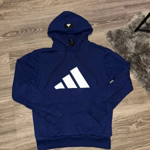 Blå hoodie från Adidas - Säljer en snygg blå hoodie från Adidas med stor vit logga på framsidan. Hoodien har en klassisk design med justerbar huva och en stor magficka. Perfekt för en sportig look eller avslappnade dagar. 🌟