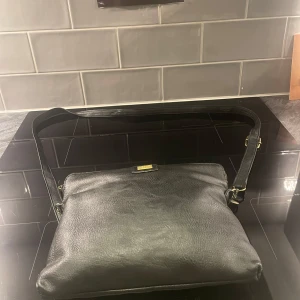 Svart dam väska från pipols bazaar - snygg svart axelväska äkta skinn från pipols bazaar. väskan har en justerbar axelrem och en praktisk dragkedja på baksidan. perfekt för att bära dina dagliga nödvändigheter med stil.