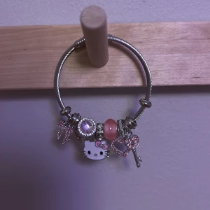 Charmarmband med bland annat Hello Kitty, nyckel,rosett  - Charmigt armband med flera hängen, inklusive en nyckel, hjärta och Hello Kitty. Armbandet har en metallisk look med inslag av rosa och silver. Perfekt för att ge en personlig touch till din stil.
