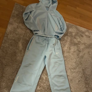 Ljusblå hoodie från Nike NOCTA - Säljer en stilren ljusblå hoodie från Nike NOCTA med dragkedja och huva. Tröjan har diskreta logotyper och praktiska fickor med dragkedja. Perfekt för en avslappnad stil.