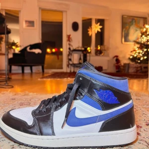 Nike Air Jordans i svart, blå och vit - Säljer ett par Nike Air Jordans i svart, blå och vit. Skorna har en klassisk hög design med snörning och en tydlig Nike-logga på sidan. Perfekt för de som gillar att variera stiler