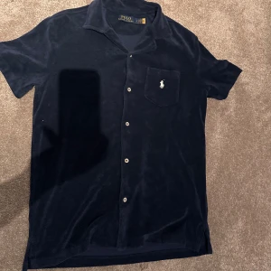 Mörkblå kortärmad skjorta från Ralph Lauren - Snygg mörkblå kortärmad skjorta från Ralph Lauren med klassisk krage och knappar framtill. Skjortan har en bröstficka med det ikoniska broderade logotypen. Perfekt för en stilren look.