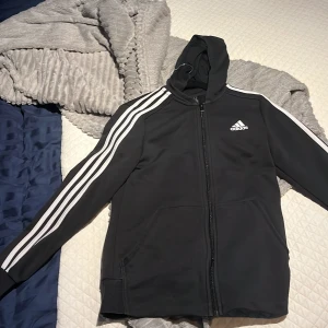 Svart huvtröja från Adidas - Säljer en svart huvtröja från Adidas med klassiska vita ränder längs ärmarna och dragkedja framtill. Tröjan har en justerbar huva och två fickor. Perfekt för en sportig look eller avslappnade dagar.