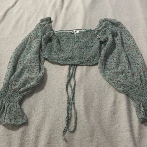 Blommig grön blus från Chiquelle - Säljer en söt blommig blus från Chiquelle i storlek medium. Den är grön med små blommor i rött och svart. Blusen har långa puffärmar och en smockad kropp med knytband framtill. Perfekt för en somrig look! 🌸