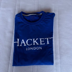 Blå t-shirt från Hackett London - Snygg blå t-shirt från Hackett London med vit logga på bröstet. Perfekt för en stilren och avslappnad look. T-shirten har en klassisk rund hals och långa armar