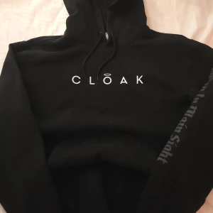 Svart hoodie från Cloak - Snygg svart hoodie från bl.a Markipliers märke Cloak med texten 'Cloak' på framsidan och 'Not That Bad' med vingar på baksidan. På armen står det "Hidden in plain sight" Den har en praktisk magficka och justerbar huva. Hoodien är uv responsive i solen och det vita på hoodie blir till en söt ljus rosa!!