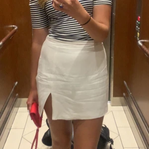 Vit kortkjol med slits - Säljer en stilren vit kortkjol med en snygg slits framtill. Kjolen har en enkel och elegant design som passar perfekt för varma dagar. Den är lätt att matcha med olika toppar och accessoarer.