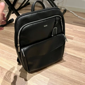 Svart ryggsäck från Tumi - Säljer en stilren och praktisk svart ryggsäck från Tumi. Den är tillverkad i skinn och har flera dragkedjeförsedda fack för enkel organisering. Perfekt för både vardag och resor!
