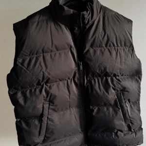 Svart dunväst från Weekday - Säljer en snygg svart dunväst från Weekday i storlek L. Västen är perfekt för höst och vinter med sitt varma och puffiga material. Den har en dragkedja framtill och är tillverkad av återvunnen polyester. 