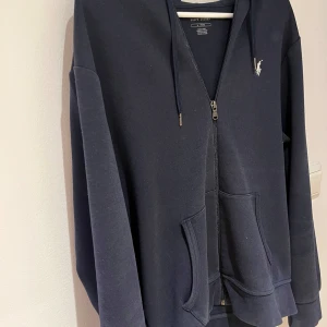 Mörkblå hoodie från Ralph Lauren - Säljer en snygg och bekväm mörkblå hoodie från Ralph Lauren. Den har en dragkedja framtill och en klassisk logga på bröstet. Perfekt för kyliga dagar eller som ett stilrent lager under jackan. Den har långa ärmar och en praktisk huva. Öppen för byten