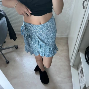 Blommig blå omlottkjol - Säljer en superfin blå omlottkjol med små vita blommor. Kjolen har en söt volangdetalj längst ner och knyts i sidan för en perfekt passform. Perfekt för sommardagar eller en mysig vårutflykt! 💙