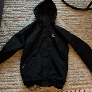 Svart vindjacka från Haglöfs - Säljer en svart vindjacka från Haglöfs i storlek M. Jackan har en praktisk huva och dragkedja framtill. Perfekt för blåsiga dagar och utomhusaktiviteter. Den är i bra skick knappt använd