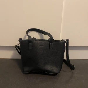 Svart axelväska från Don Donna - Snygg svart axelväska från Don Donna i skinn. Väskan har en praktisk dragkedja och justerbar axelrem. Perfekt för att bära dina dagliga nödvändigheter med stil.