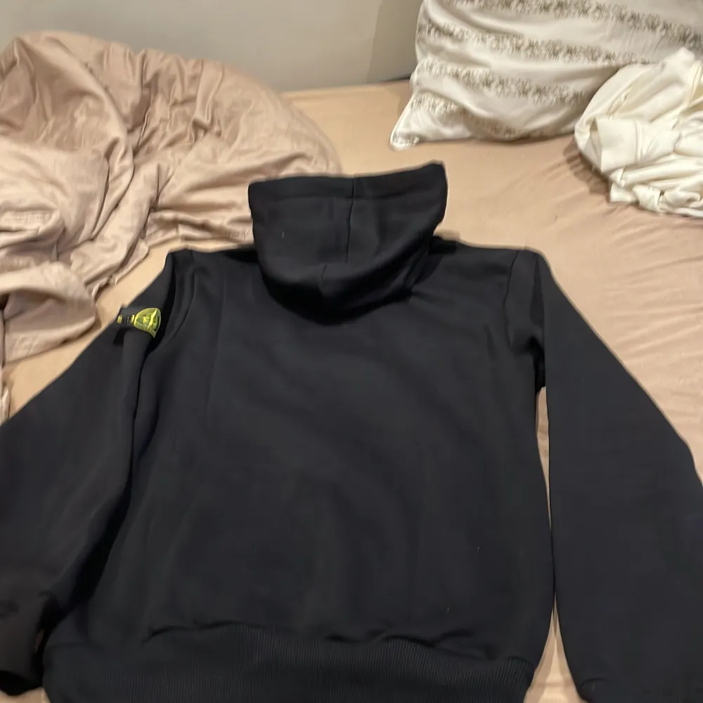 Säljer en svart hoodie från Stone Island med det ikoniska kompassmärket på bröstet och en avtagbar patch på ärmen. Tröjan har en klassisk huva med dragsko och ribbade muddar vid ärmslut och nederkant. Perfekt för en stilren och avslappnad look.. Neuletakit & Villapaidat.