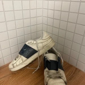 Valentino sneakers med blå detalj - Snygga Valentino sneakers i vitt med en kontrasterande blå detalj över snörningen. Skorna har en klassisk design med snörning och är tillverkade i skinn. Perfekta för en stilren look.