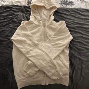 Beige hoodie/kofta i ekologisk bomull - Mysig beige hoodie i ekologisk bomull med dragkedja och huva. Perfekt för en avslappnad stil. Den har långa ärmar och en klassisk passform.