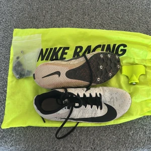 Nike Racing Spikskor - Säljer ett par Nike Racing spikskor i vitt med svarta detaljer. Skorna har snörning och en mönstrad ovandel med svarta Nike-loggor. Perfekta för löpning med sina utbytbara spikar på sulan. Kommer med en neongul förvaringspåse. Samt så finns det extra skruvar.