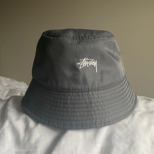 Stussy Bucket hat   - Snygg bucket hat från Stussy med broderad logga framtill. Perfekt för en avslappnad stil. Tillverkad i ett lätt och bekvämt material.