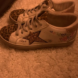 Vita sneakers med leopardmönster och glittrig stjärna - Unika vita sneakers med leopardmönstrade detaljer och en glittrig stjärna på sidan. Skorna har snörning och är dekorerade med små hjärtan i olika färger. Perfekta för att ge din outfit en extra touch!