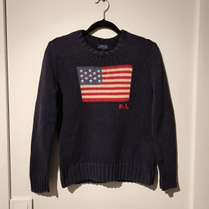 Mörkblå stickad tröja från Ralph Lauren - Säljer en snygg mörkblå stickad tröja från Ralph Lauren till barn med ett amerikanskt flaggmotiv på framsidan. Tröjan har långa ärmar och ribbstickade kanter. Perfekt för en stilren look! Storlek M i barnstorlek.