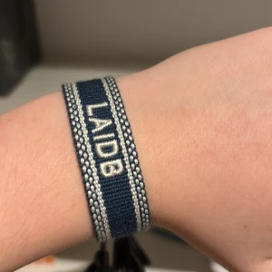 Blått armband med text - Snyggt blått armband med texten 'LAIDB' i vitt. Armbandet har en enkel design med tofsar i ändarna för justering. Perfekt för att ge en avslappnad stil.