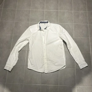 Vit långärmad skjorta - Snygg vit långärmad skjorta från H&M med klassisk krage och knappar framtill. Perfekt för en stilren look. Passar bra till både jeans och kostymbyxor. Skjortan är i 100% bomull och är i storlek: 13-14 (164cm). Varsågoda att skriva vid funderingar!