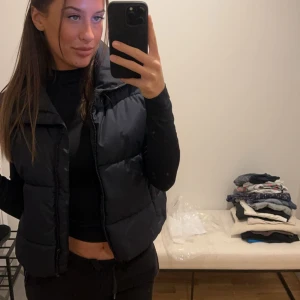 Svart dunväst - Snygg svart dunväst perfekt för kyligare dagar. Västen har en dragkedja framtill. Den är stilren och lätt att matcha med olika outfits.